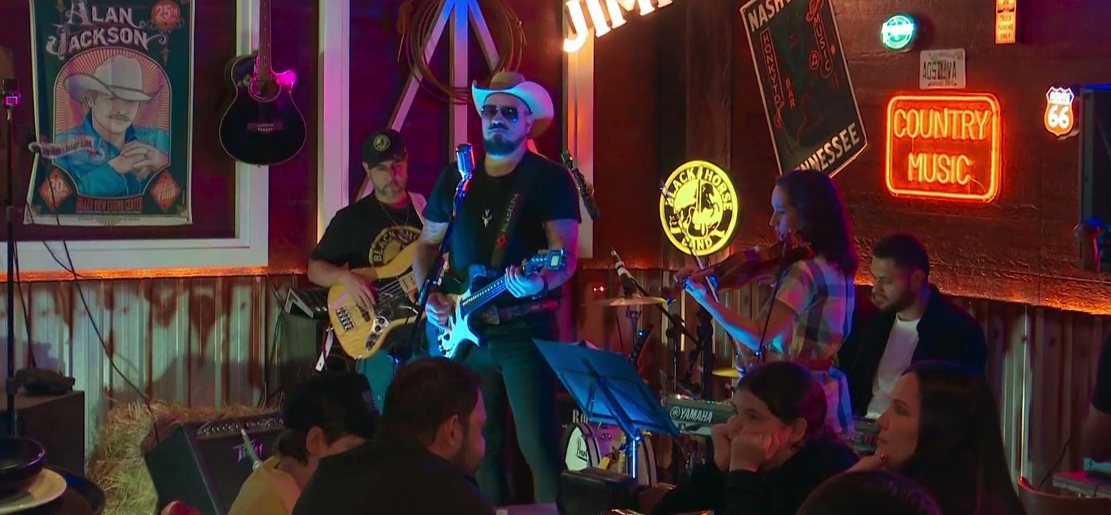 Black Horse Band completa no palco do Jimmy's: baixo, guitarra, violino e teclado, com pôster do Alan Jackson e neons ao fundo