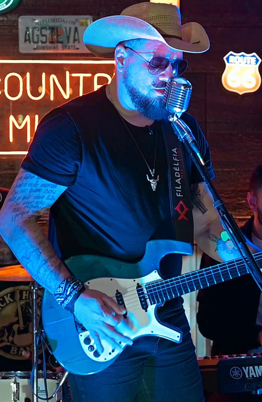 Marcelo Sá de chapéu e óculos cantando e tocando guitarra no Jimmy's Bar, com neon Country Music e placa Route 66 ao fundo