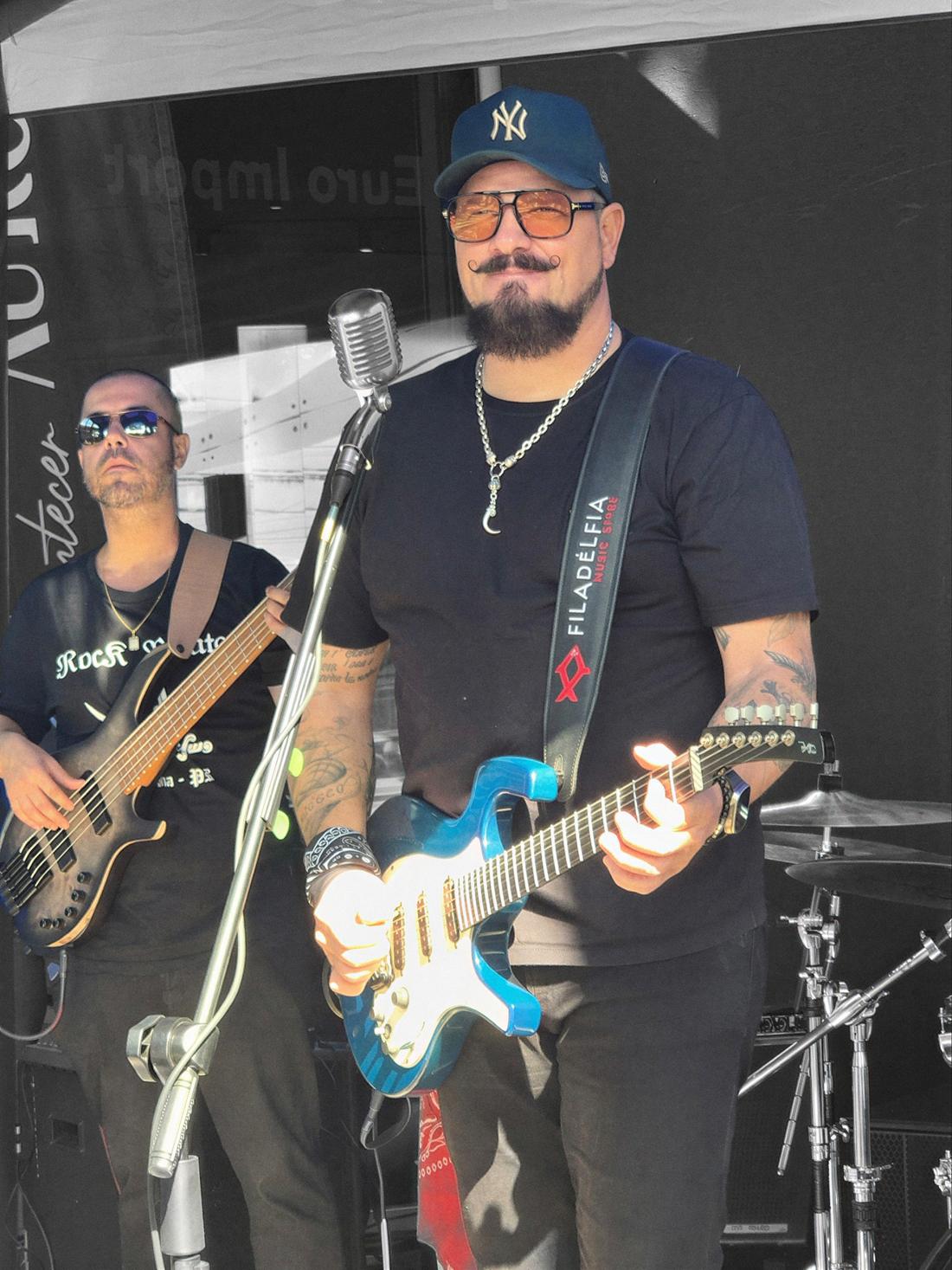 Marcelo Sá de boné e óculos tocando guitarra azul com a banda num evento durante o dia