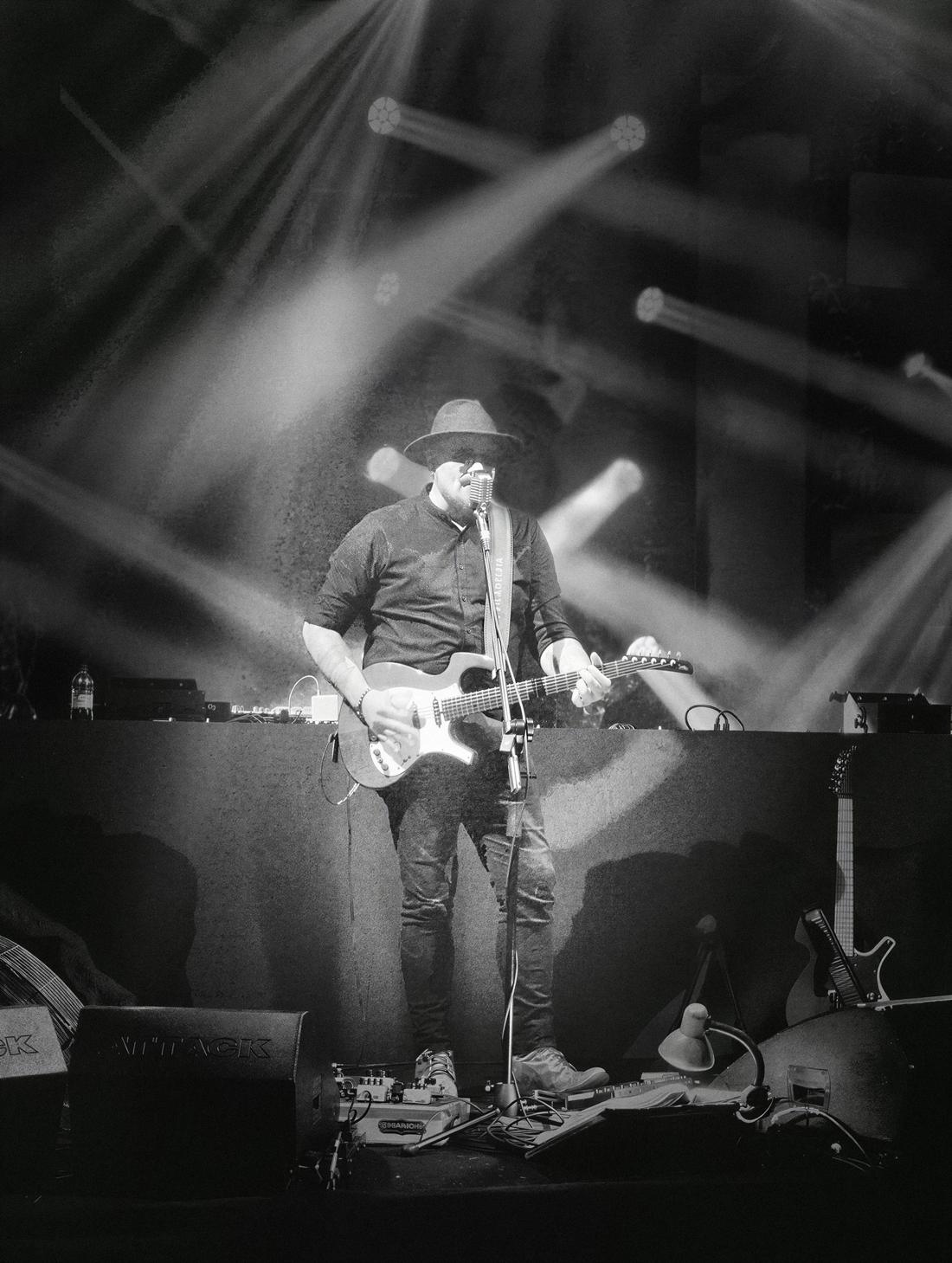 Foto em preto e branco de Marcelo Sá no palco com guitarra, cercado por fachos de luz
