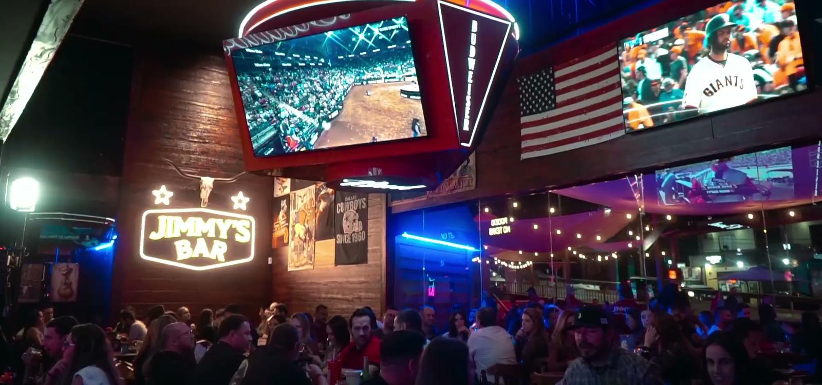 Salão do Jimmy's Bar lotado à noite, com telões, letreiro em neon e bandeira americana